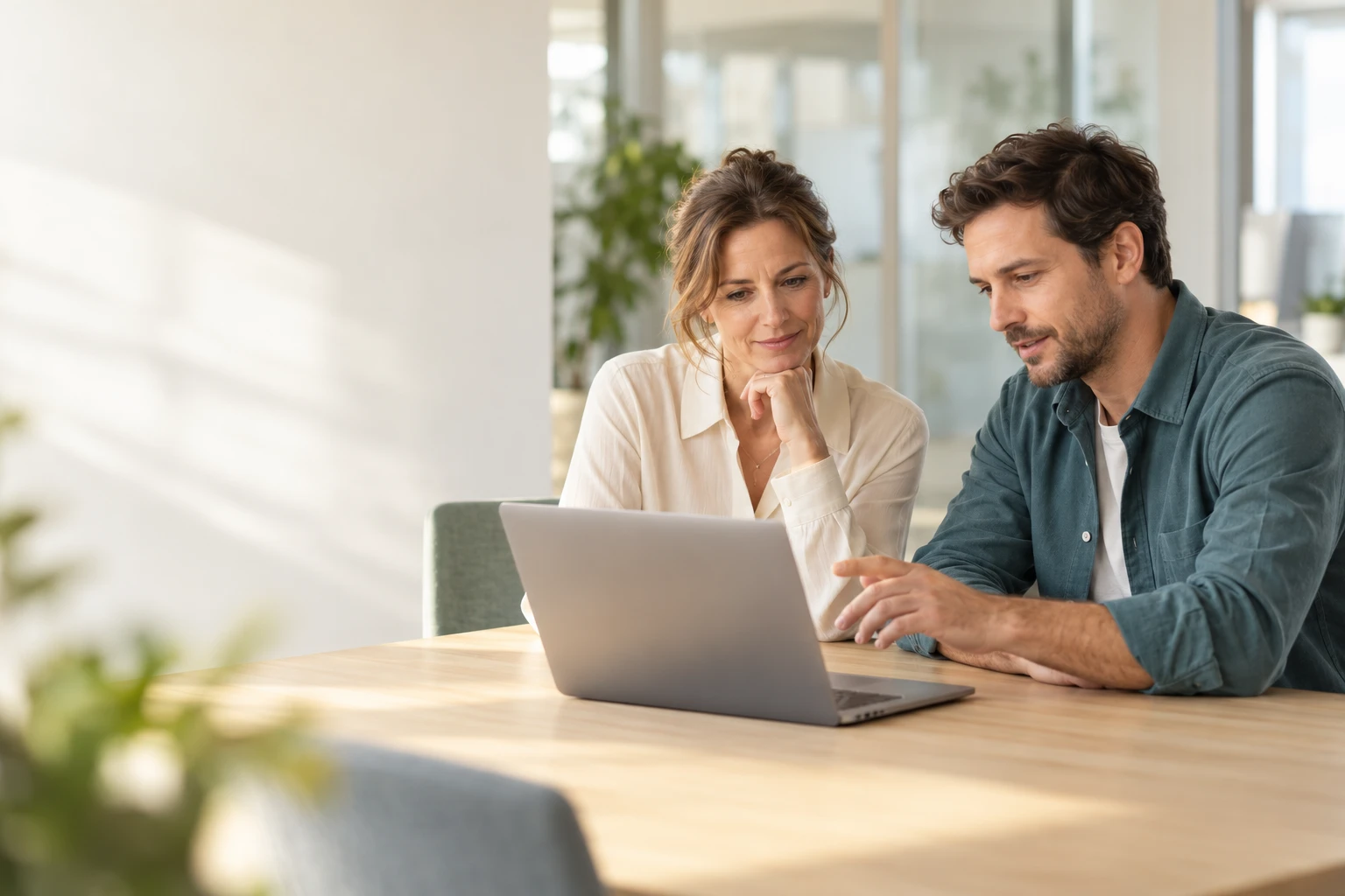 A woman and a man sit at a table in a bright office and discuss something on a laptop. (AI-generated)